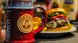 A close-up of a red ceramic mug with "The Barn - Martha's Vineyard" logo featuring an owl, placed on a table. In the slightly blurred background, a cheeseburger with lettuce, tomato, and onion sits on a plate with a side of fries.
